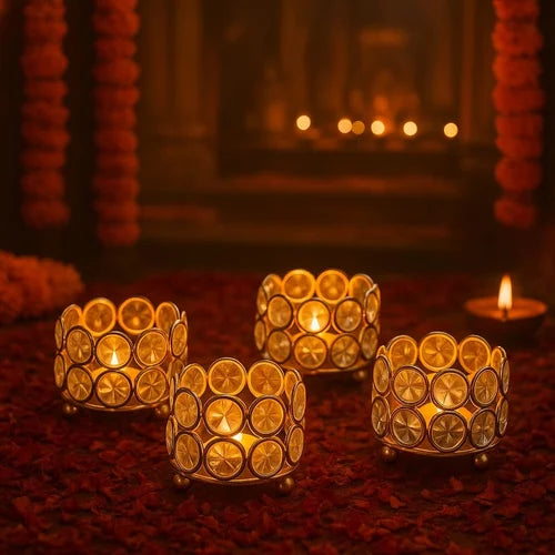 Four gold-toned metal tealight candle holders with circular cutout patterns, each lit with a small tealight candle, arranged on a bed of red flower petals in a warm, festive setting.