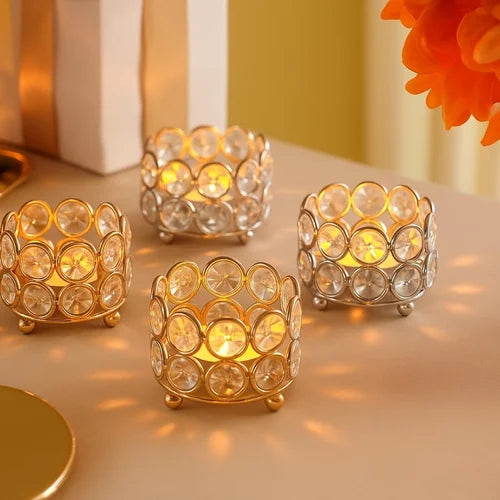 Four decorative tealight candle holders with circular clear crystal-like discs on gold and silver bases, lit with warm yellow candles and placed on a beige tabletop.