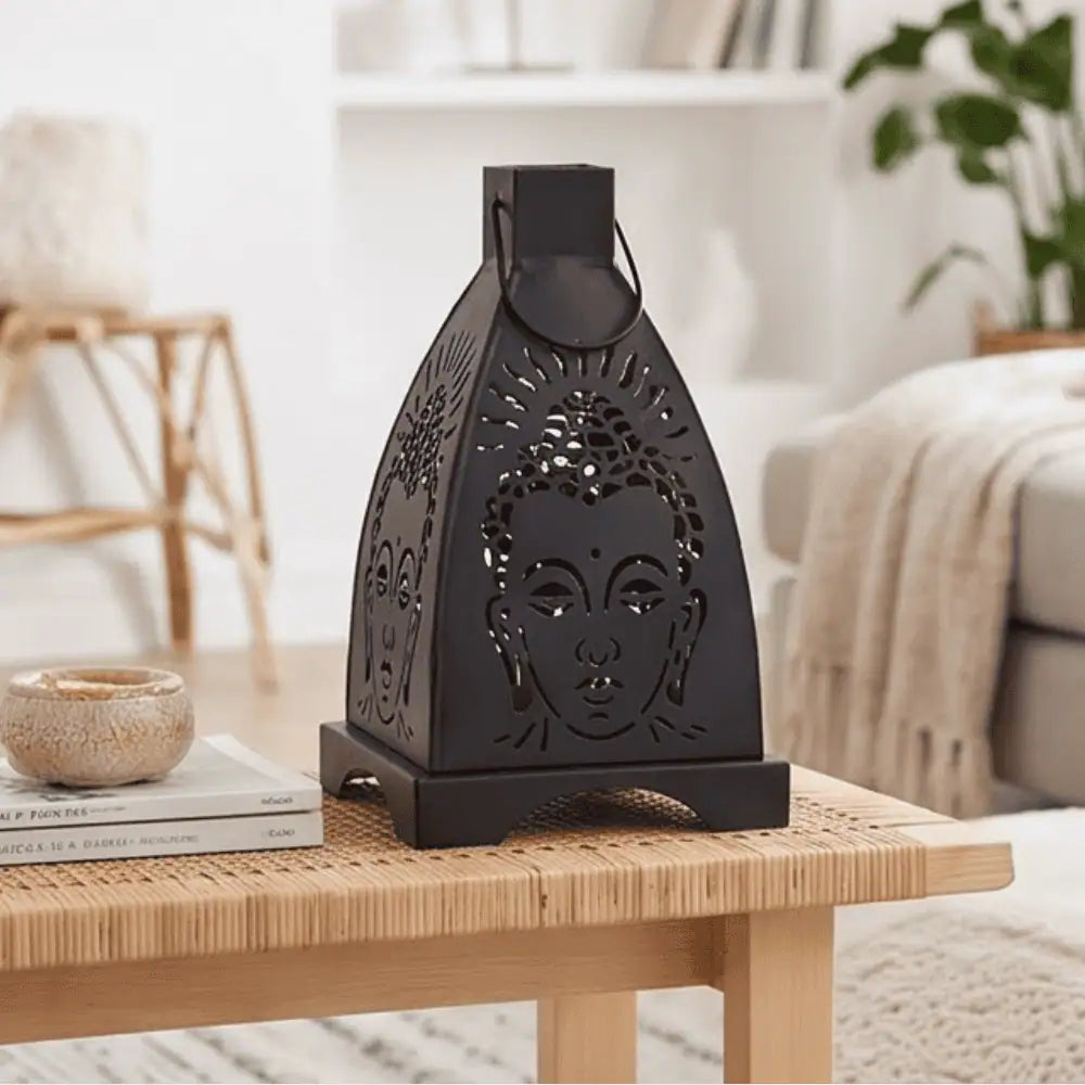A black metal Buddha head lantern with laser-cut details, displayed on a light wood woven table in a bright, modern living room setting.
