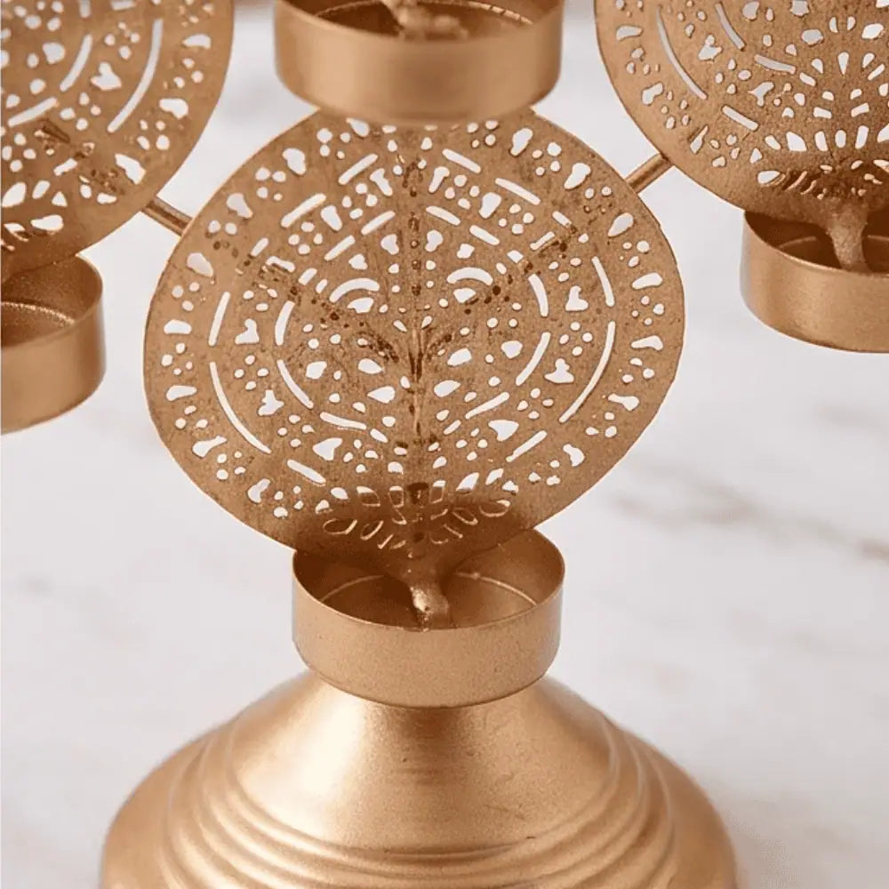 A close-up of a gold-toned metal candle holder with intricate circular cutout patterns and small tea light cups, set against a light marble background.