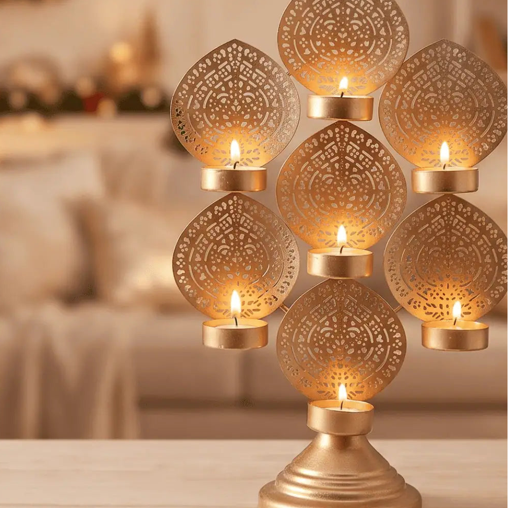 A gold-toned metal candle holder with seven ornate cutout leaf-shaped panels, each holding a small tealight candle, displayed on a tabletop in a softly blurred indoor setting.