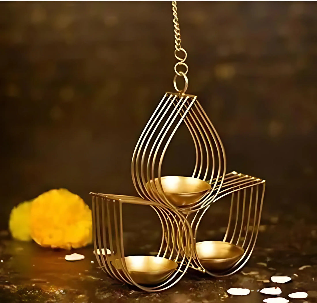 A gold-toned hanging metal diya holder with three tiers and small cup-shaped candle holders, shown on a dark textured surface with a marigold and scattered petals.