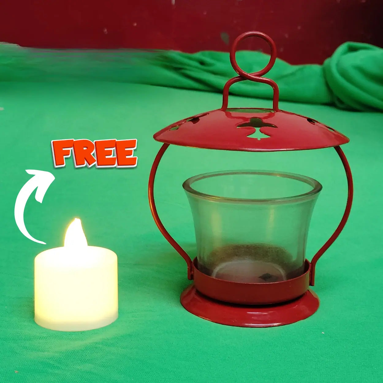 A red metal tea light lantern with a clear glass cup inside, shown on a green fabric background next to a lit white tea light candle. The word 'FREE' appears in orange text with a white arrow pointing toward the lantern.